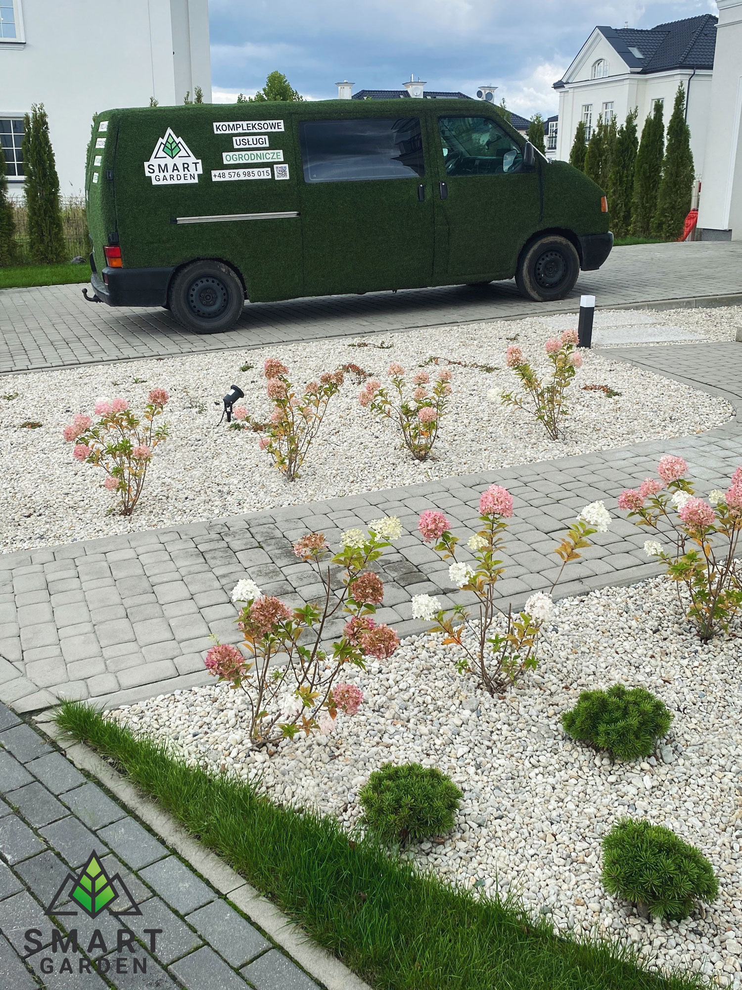 Ogród z kwitnącymi hortensjami na białym żwirze, obok zaparkowany samochód obklejony sztuczną trawą z logo 'Smart Garden'.