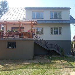 Patryk Mućka - Dwukondygnacyjny dom z jasną elewacją, drewnianą pergolą na balkonie i schodami zewnętrznymi, widoczne donice z kwiatami na balustradzie, dolna część elewacji w kolorze szarym.