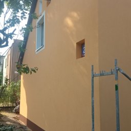 Patryk Mućka - Ściana budynku po nałożeniu tynku w kolorze piaskowym z brązowym cokołem, widoczny fragment rusztowania i tabliczka z numerem domu w małej wnęce.