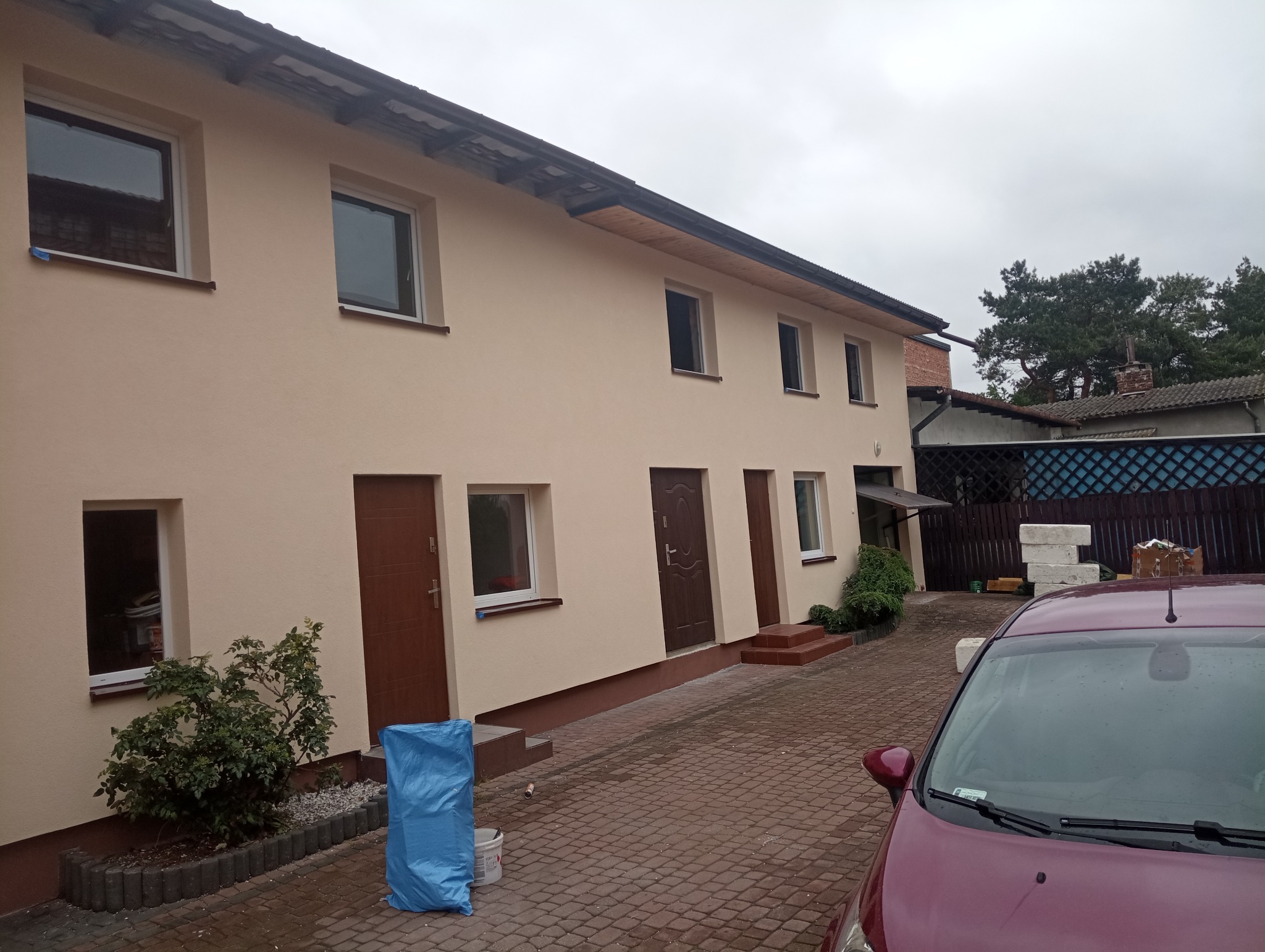Dwukondygnacyjny dom z beżową elewacją i brązowymi drzwiami, widok z zewnątrz, z widocznym fragmentem samochodu na pierwszym planie i niebieskim workiem zakrywającym wiadro farby.
