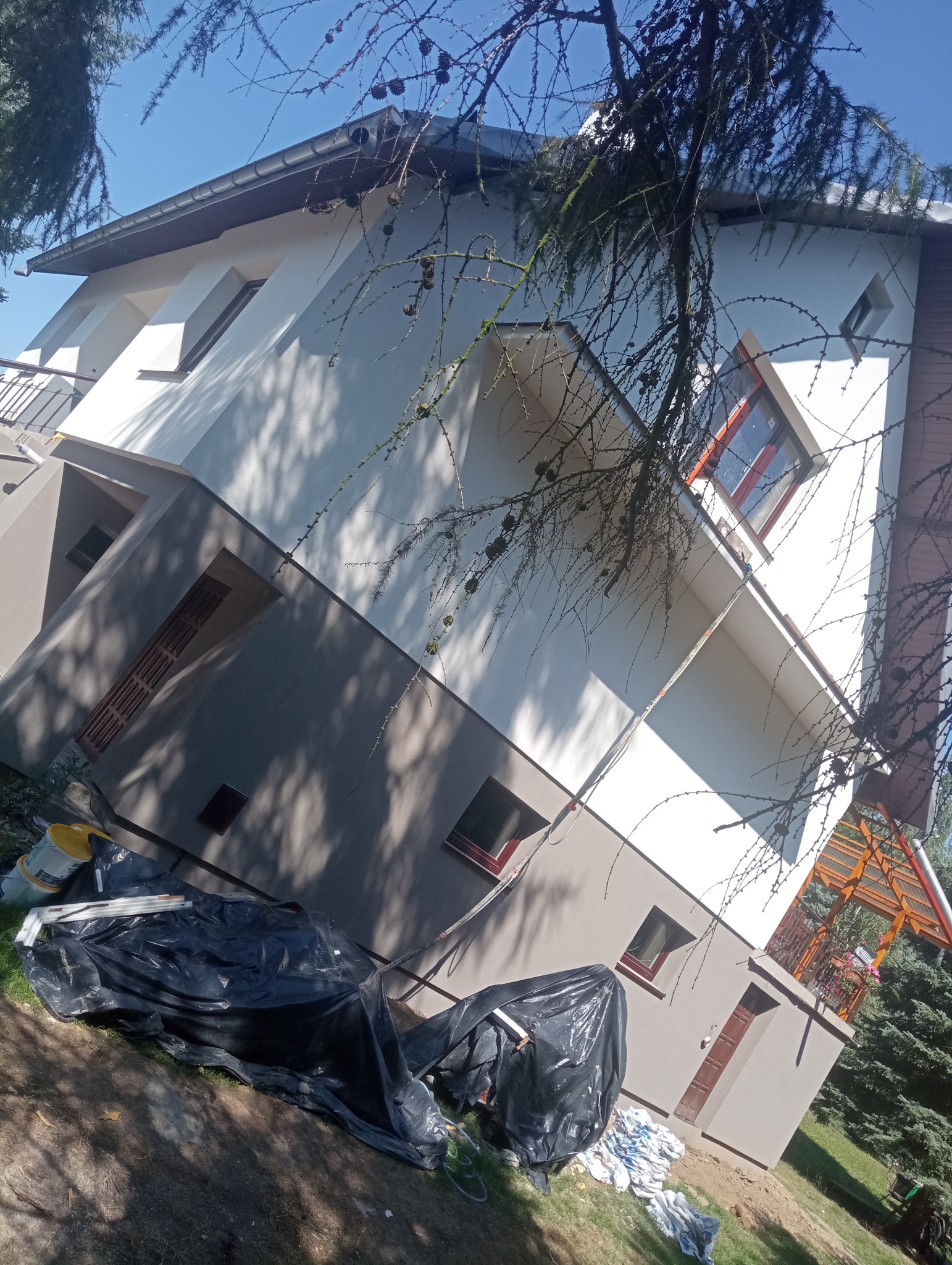 Dwukolorowa fasada domu po remoncie, z widocznymi detalami wykończeniowymi, zabezpieczona folią budowlaną, z fragmentem drzewa iglastego w kadrze.