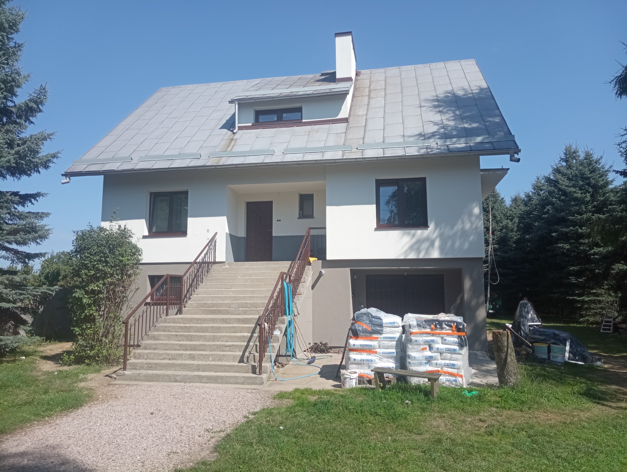 Dwupiętrowy dom z szarą dachówką i białymi ścianami, z betonowymi schodami prowadzącymi do wejścia, z materiałami budowlanymi obok schodów i rozłożonymi narzędziami malarskimi w tle na trawie.