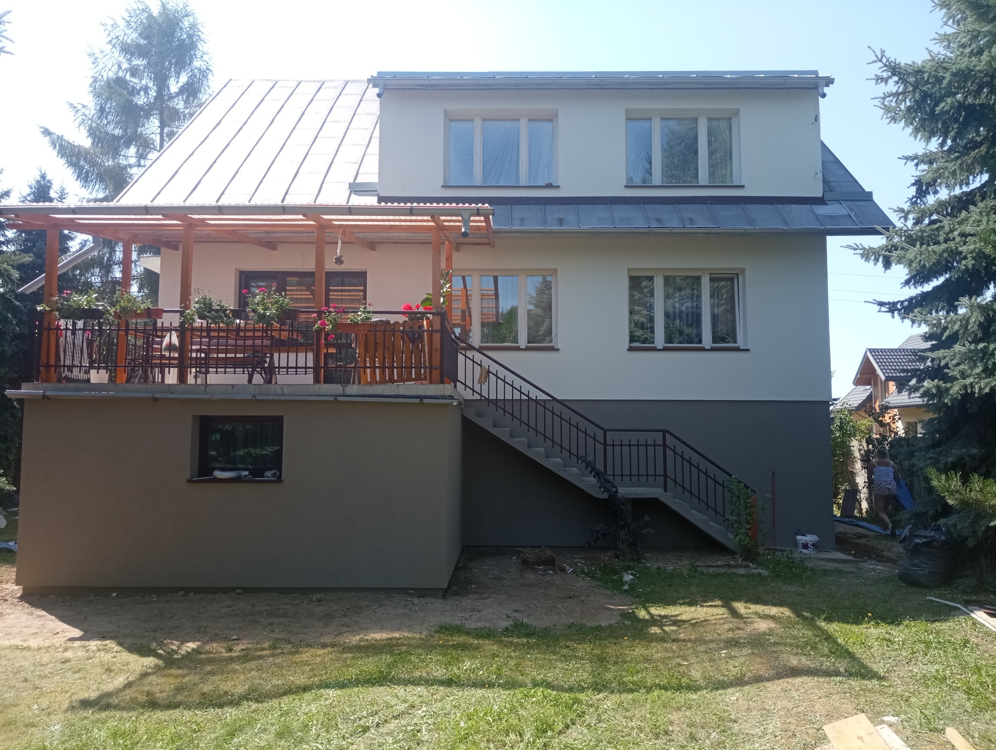 Dwukondygnacyjny dom z jasną elewacją, drewnianą pergolą na balkonie i schodami zewnętrznymi, widoczne donice z kwiatami na balustradzie, dolna część elewacji w kolorze szarym.