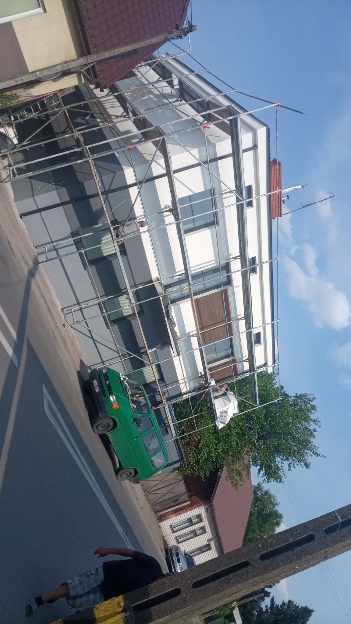 Dom w trakcie prac elewacyjnych, widoczne rusztowania, częściowo pomalowana fasada na biało i szaro, na ulicy zaparkowany zielony dostawczy samochód, na rusztowaniu pracownik w jasnej odzieży.