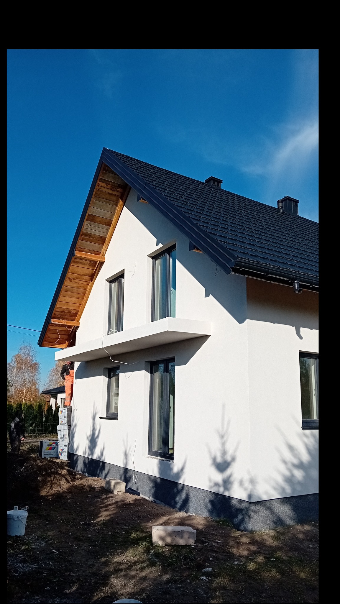 Nowo wykończona elewacja domu jednorodzinnego z czarną dachówką, widoczne elementy drewniane pod dachem, pracownik w pomarańczowym kombinezonie na rusztowaniu oraz materiały budowlane w pobliżu.