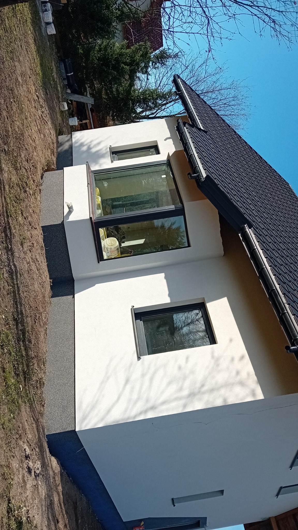 Nowoczesny dom z białymi ścianami i dużym oknem narożnym, widok z dołu, ciemna dachówka, błękitne niebo w tle.