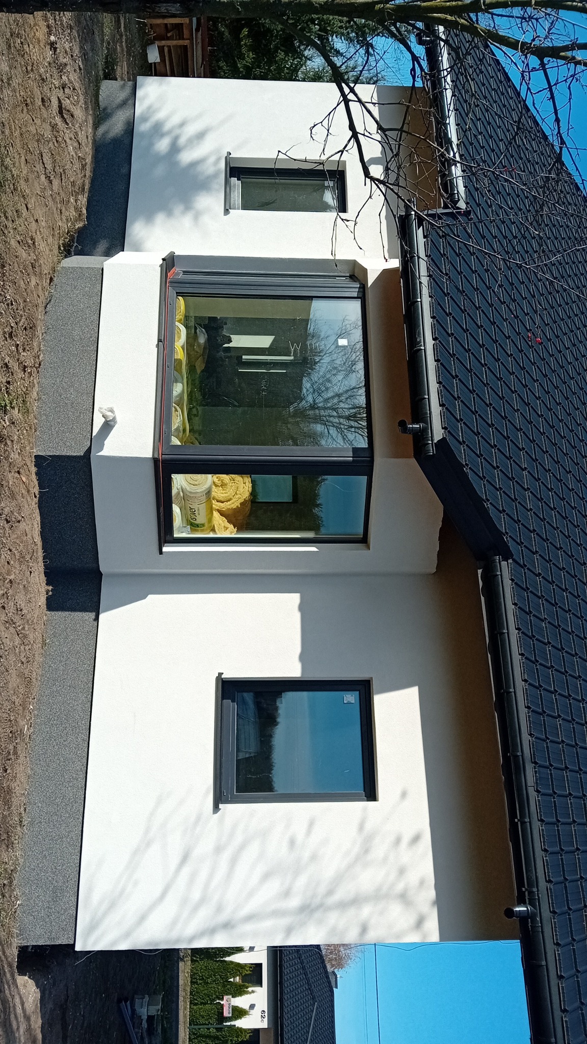 Nowoczesny dom z oknem wykuszowym, widoczne rolki wełny mineralnej wewnątrz, ściana pokryta gładkim, białym tynkiem, czarna dachówka i orynnowanie.