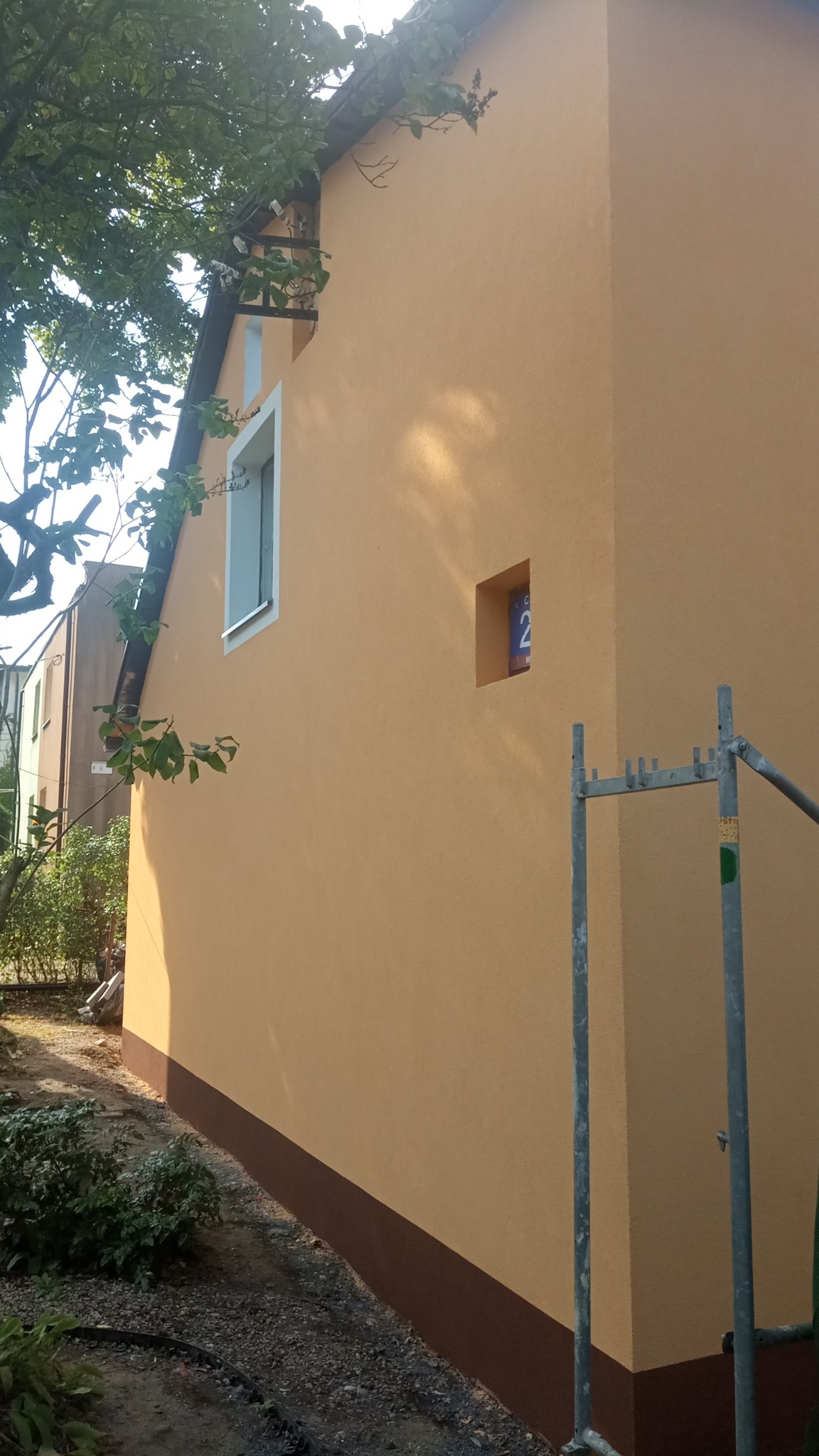 Ściana budynku po nałożeniu tynku w kolorze piaskowym z brązowym cokołem, widoczny fragment rusztowania i tabliczka z numerem domu w małej wnęce.