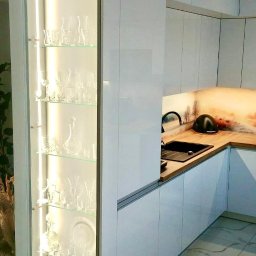 PSH USŁUGI SPÓŁKA Z OGRANICZONĄ ODPOWIEDZIALNOŚCIĄ - Biała zabudowa kuchenna z przeszkloną witryną podświetloną LED, eksponującą kolekcję szklanych naczyń, z czarnym zlewem i baterią na drewnianym blacie z motywem pejzażu.