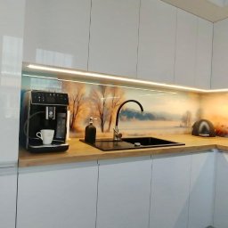 PSH USŁUGI SPÓŁKA Z OGRANICZONĄ ODPOWIEDZIALNOŚCIĄ - Biała, nowoczesna zabudowa kuchenna z blatem imitującym drewno, podświetleniem LED i panelem dekoracyjnym z motywem zimowego krajobrazu, ekspres do kawy z filiżanką, dozownik na mydło i toster.