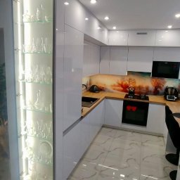 PSH USŁUGI SPÓŁKA Z OGRANICZONĄ ODPOWIEDZIALNOŚCIĄ - Biała, narożna kuchnia z drewnianym blatem i szklanym panelem z motywem drzew, oświetlona witryna z szkłem i porcelaną, marmurowa podłoga.