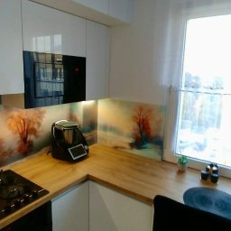 PSH USŁUGI SPÓŁKA Z OGRANICZONĄ ODPOWIEDZIALNOŚCIĄ - Nowoczesna kuchnia z białymi, błyszczącymi frontami, drewnianym blatem i panelem szklanym z motywem drzew, widoczny Thermomix i płyta grzewcza.