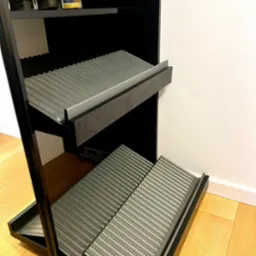 Czarna szafka na buty z wysuwanymi, żebrowanymi półkami, ustawiona na drewnianej podłodze przy białej ścianie.