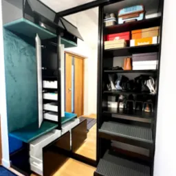 Czarna szafa wnękowa z lustrzanymi drzwiami przesuwnymi, wewnętrzne półki z pudełkami na buty i bagażem, obok siedzisko z zieloną tapicerką i otwartymi szafkami, drewniana podłoga.