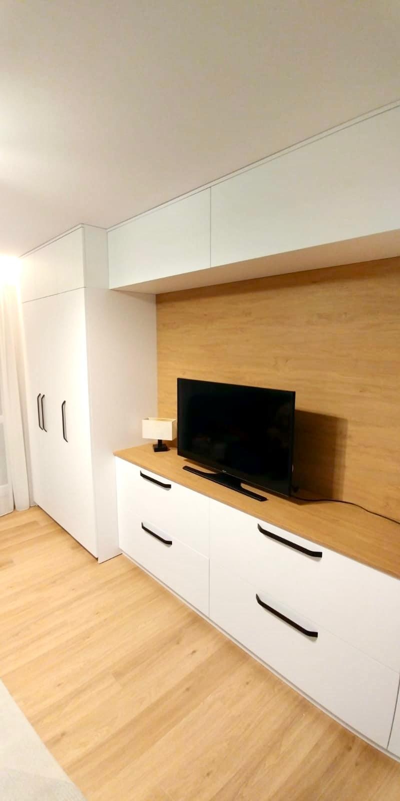 Biała meblościanka z szafą i komodą pod telewizor, z drewnianym blatem i panelem ściennym, czarne uchwyty, nowoczesny minimalizm.