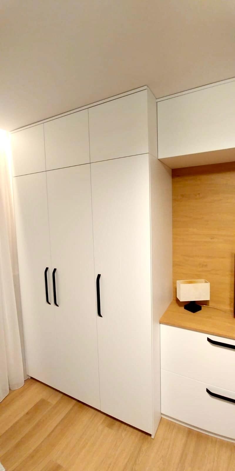 Biała szafa wnękowa z czarnymi, minimalistycznymi uchwytami, dopasowana do zabudowy, obok komoda z drewnianym blatem i lampką nocną.