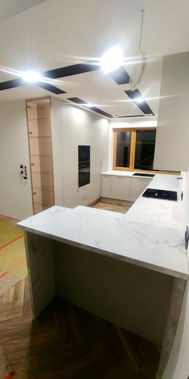 Jasna kuchnia z blatem imitującym marmur, widoczna płyta indukcyjna, zabudowa meblowa od podłogi do sufitu, oświetlenie sufitowe z czarnymi elementami dekoracyjnymi, drewniana podłoga ułożona...