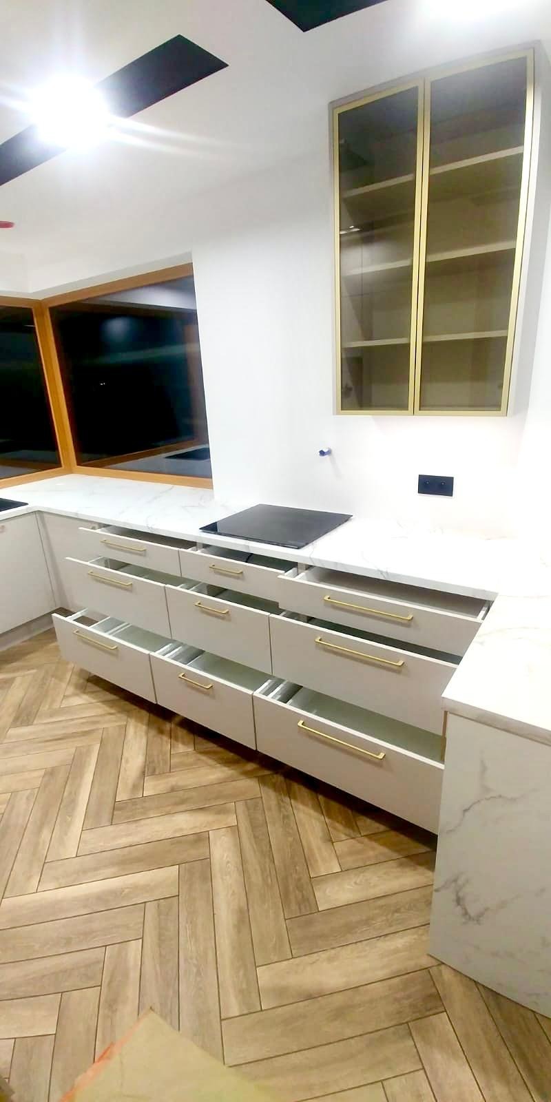 Nowoczesna kuchnia z białymi szafkami z marmurowym blatem, złotymi uchwytami i częściowo otwartymi szufladami, na podłodze parkiet w jodełkę.