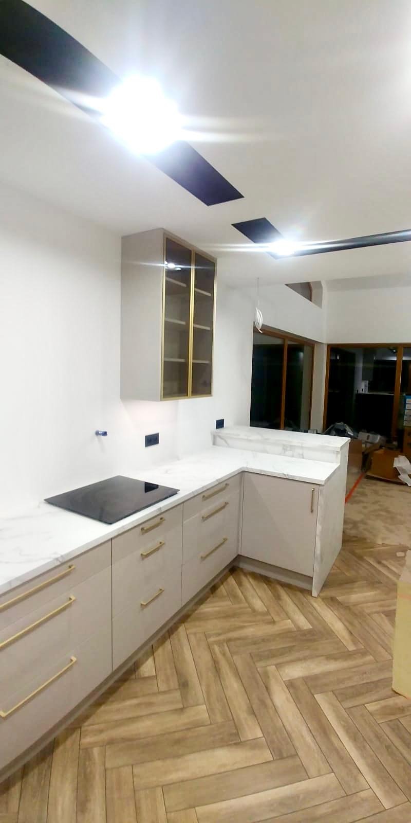 Jasna kuchnia z marmurowym blatem, złotymi uchwytami i płytą indukcyjną, widok na podłogę ułożoną w jodełkę oraz przeszklone drzwi balkonowe w tle.