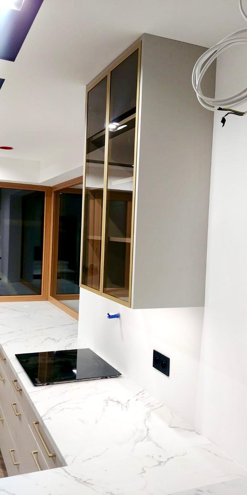 Szklana szafka wisząca z złotymi ramami nad blatem z imitacji marmuru, płyta indukcyjna w trakcie montażu, widoczne kable elektryczne.