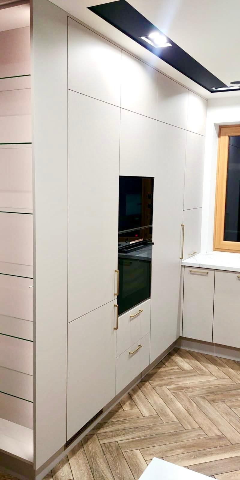 Jasna zabudowa kuchenna z piekarnikiem w zabudowie, złotymi uchwytami i szklanymi półkami po lewej stronie, na podłodze parkiet ułożony w jodełkę.