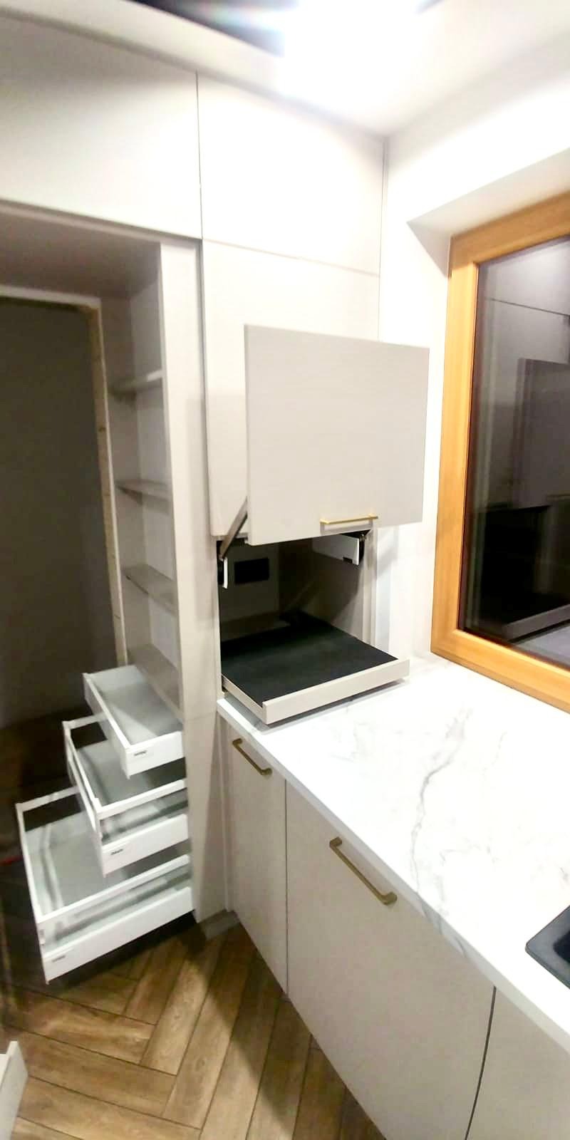 Nowoczesna kuchnia: białe szafki z wysuwanymi szufladami i półkami, marmurowy blat, złote uchwyty, drewniane okno.