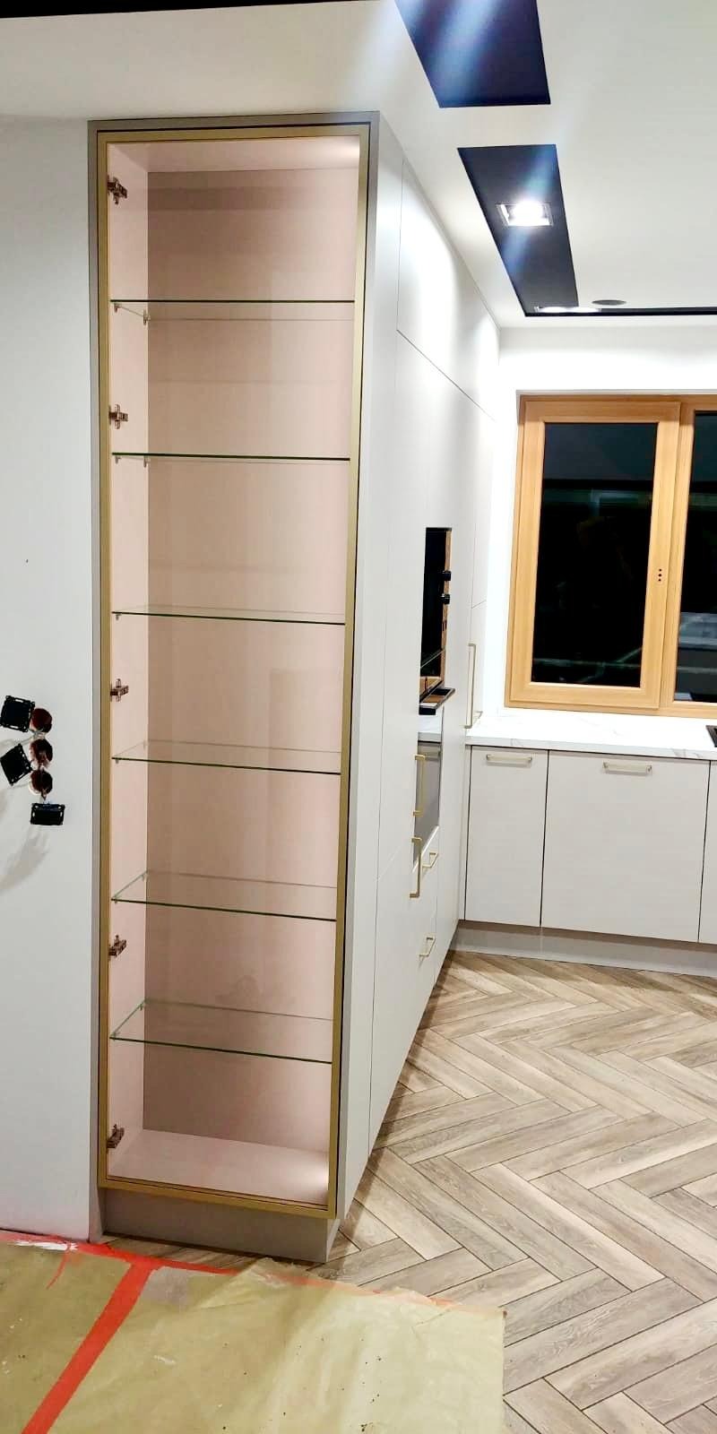 Wysoka, otwarta szafka z półkami ze szkła i złotymi obramowaniami, wkomponowana w zabudowę kuchenną z jasnymi frontami i drewnianą podłogą w jodełkę.