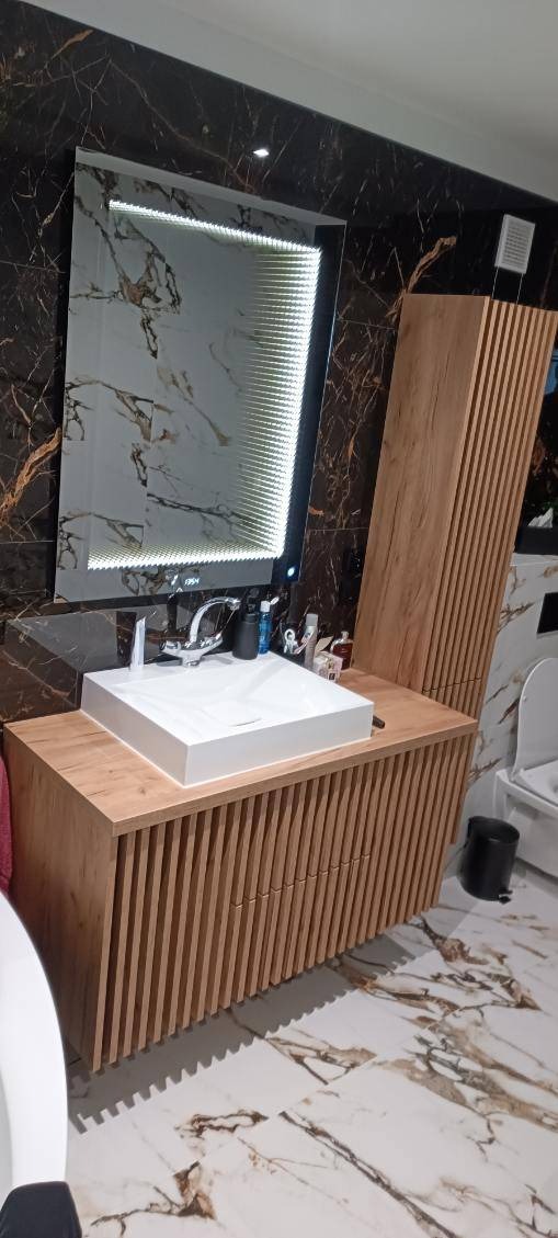 Nowoczesna łazienka z drewnianą szafką pod umywalkę z pionowymi listwami, prostokątną białą umywalką nablatową i podświetlanym lustrem z efektem nieskończoności na ścianie z czarnego marmuru.