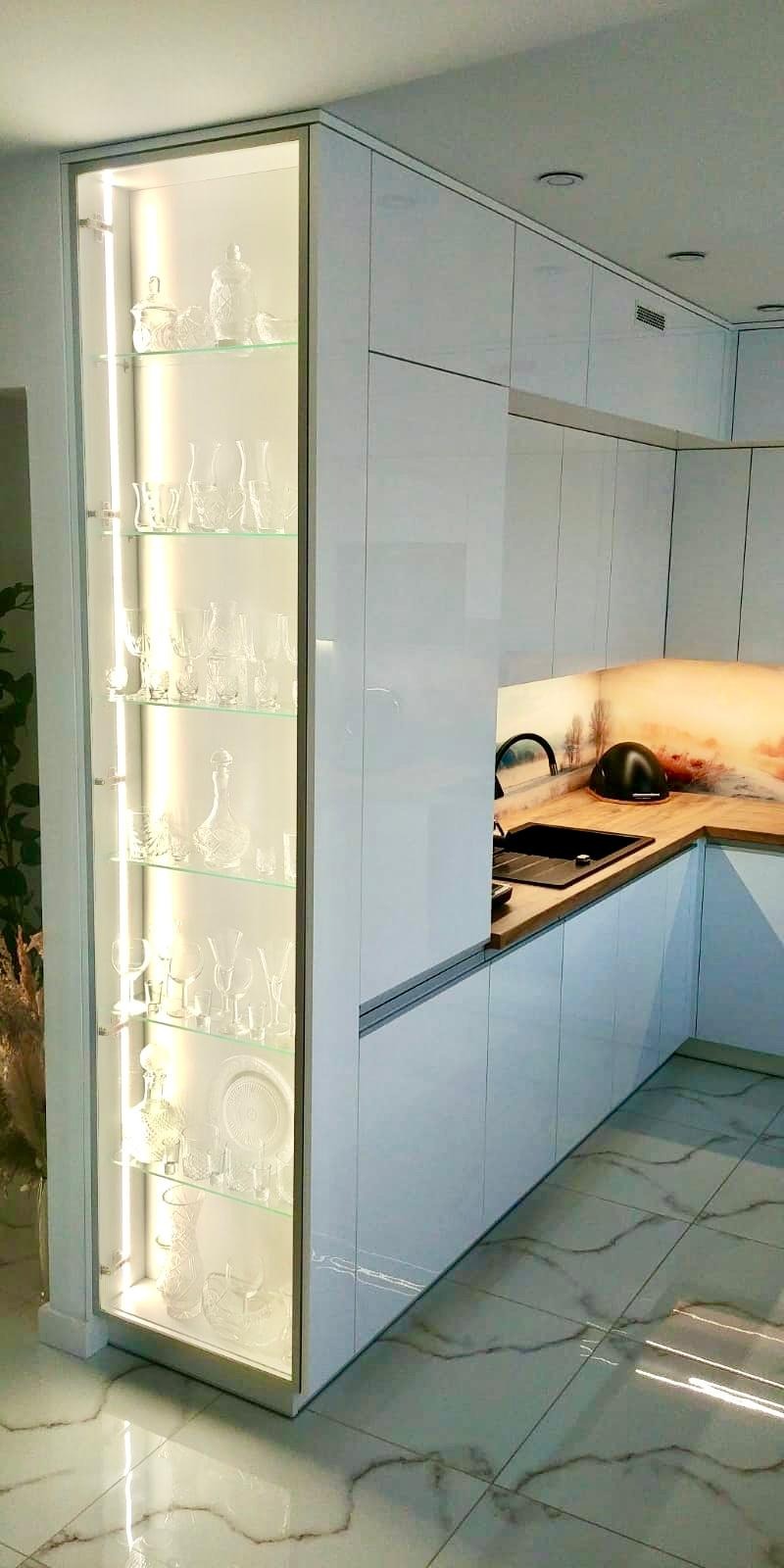 Biała zabudowa kuchenna z przeszkloną witryną podświetloną LED, eksponującą kolekcję szklanych naczyń, z czarnym zlewem i baterią na drewnianym blacie z motywem pejzażu.