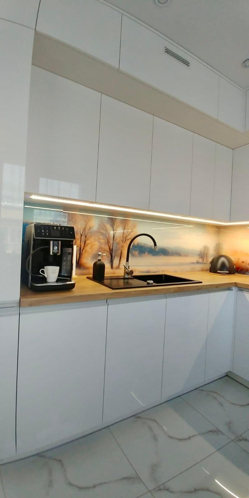 Biała, nowoczesna zabudowa kuchenna z blatem imitującym drewno, podświetleniem LED i panelem dekoracyjnym z motywem zimowego krajobrazu, ekspres do kawy z filiżanką, dozownik na mydło i toster.