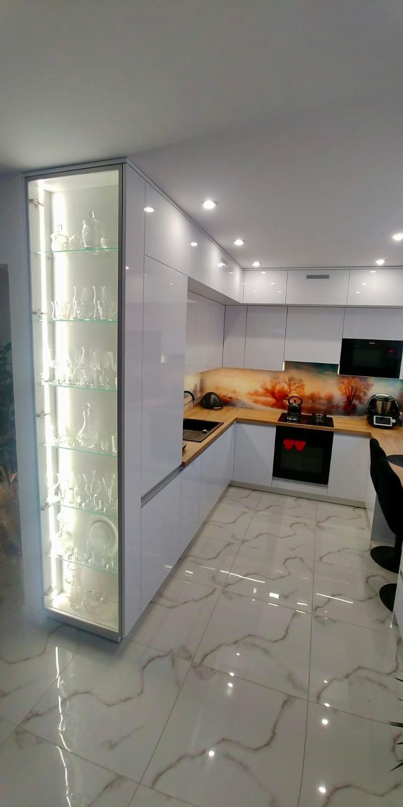 Biała, narożna kuchnia z drewnianym blatem i szklanym panelem z motywem drzew, oświetlona witryna z szkłem i porcelaną, marmurowa podłoga.