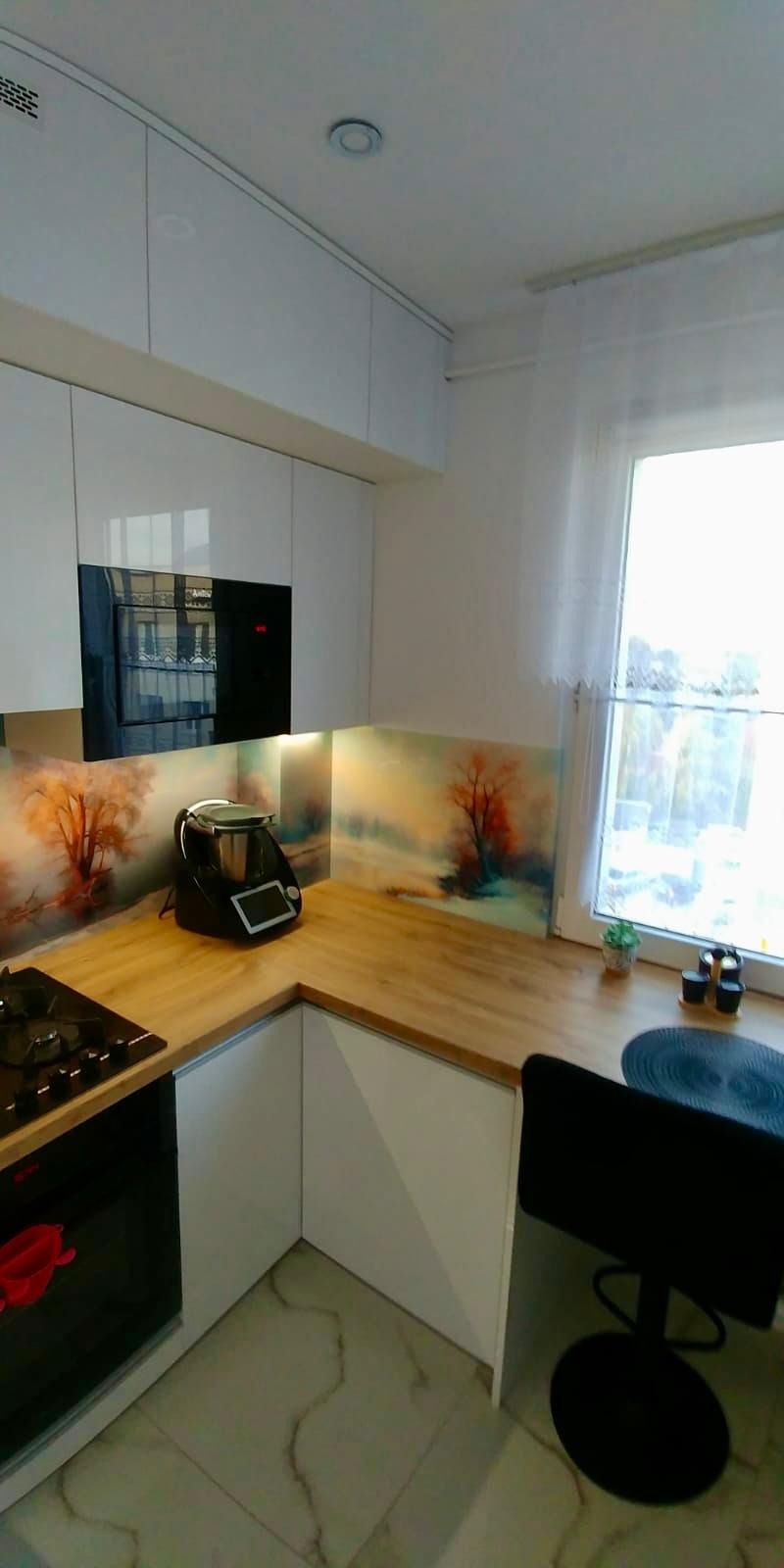 Nowoczesna kuchnia z białymi, błyszczącymi frontami, drewnianym blatem i panelem szklanym z motywem drzew, widoczny Thermomix i płyta grzewcza.