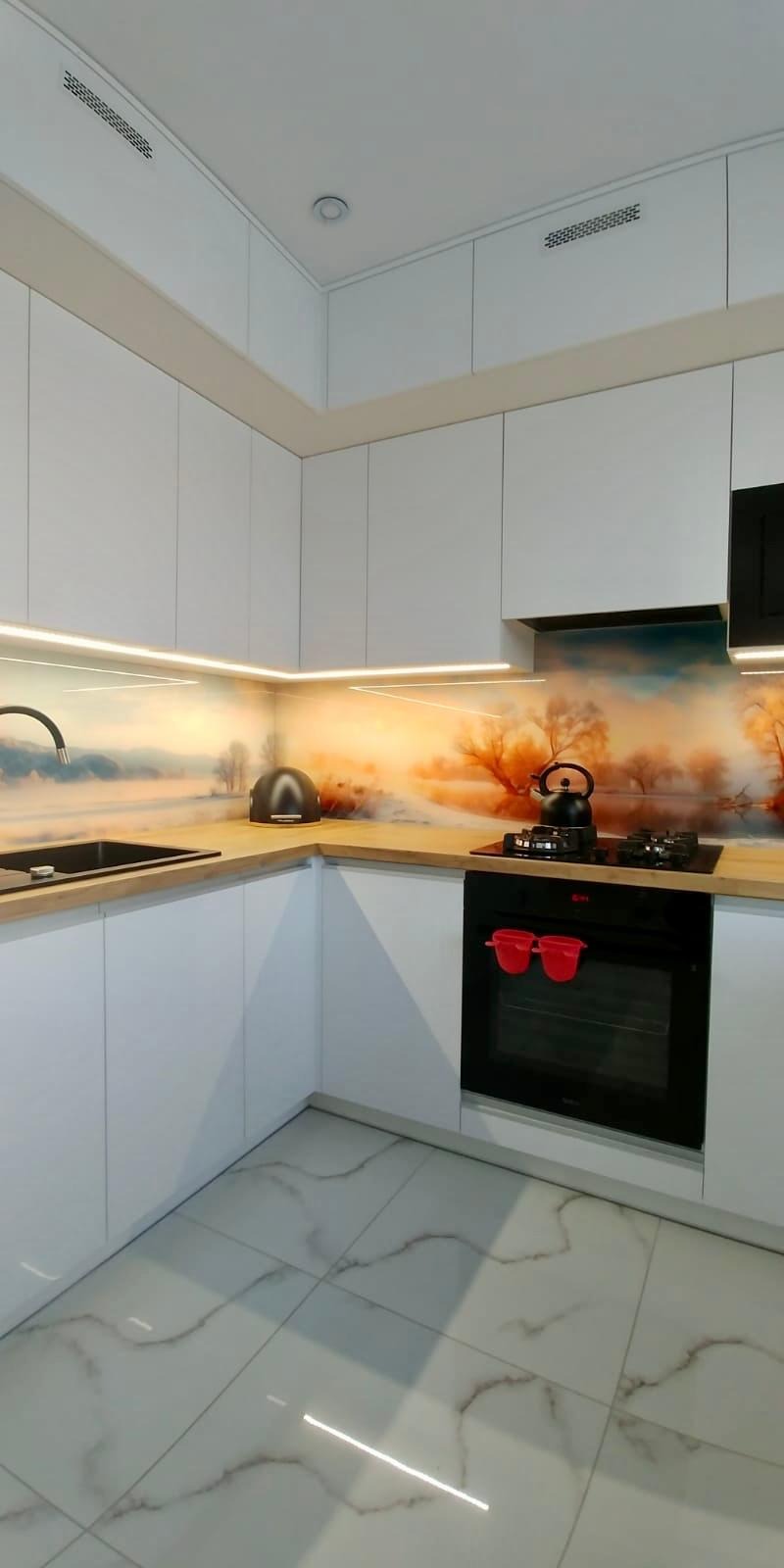 Biała kuchnia z frontami bez uchwytów, blat z drewnopodobnym dekorem, szklany panel nad blatem z motywem pejzażu, czarny piekarnik z czerwonymi uchwytami, biała marmurowa podłoga.