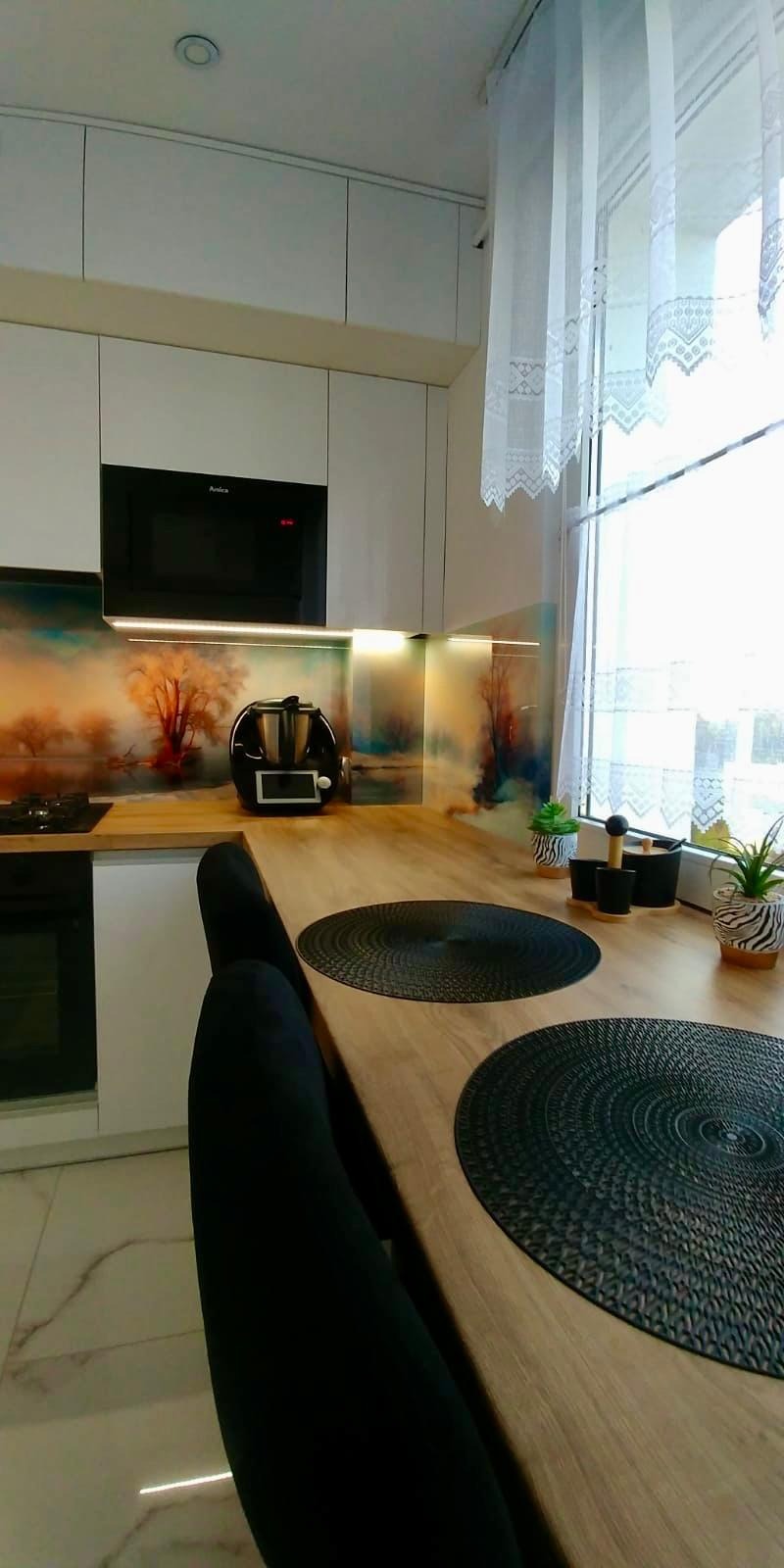 Nowoczesna biała kuchnia z blatem imitującym drewno, ozdobnym panelem szklanym z motywem leśnym i czarnym sprzętem AGD Amica, widok na okno z koronkowymi firankami.