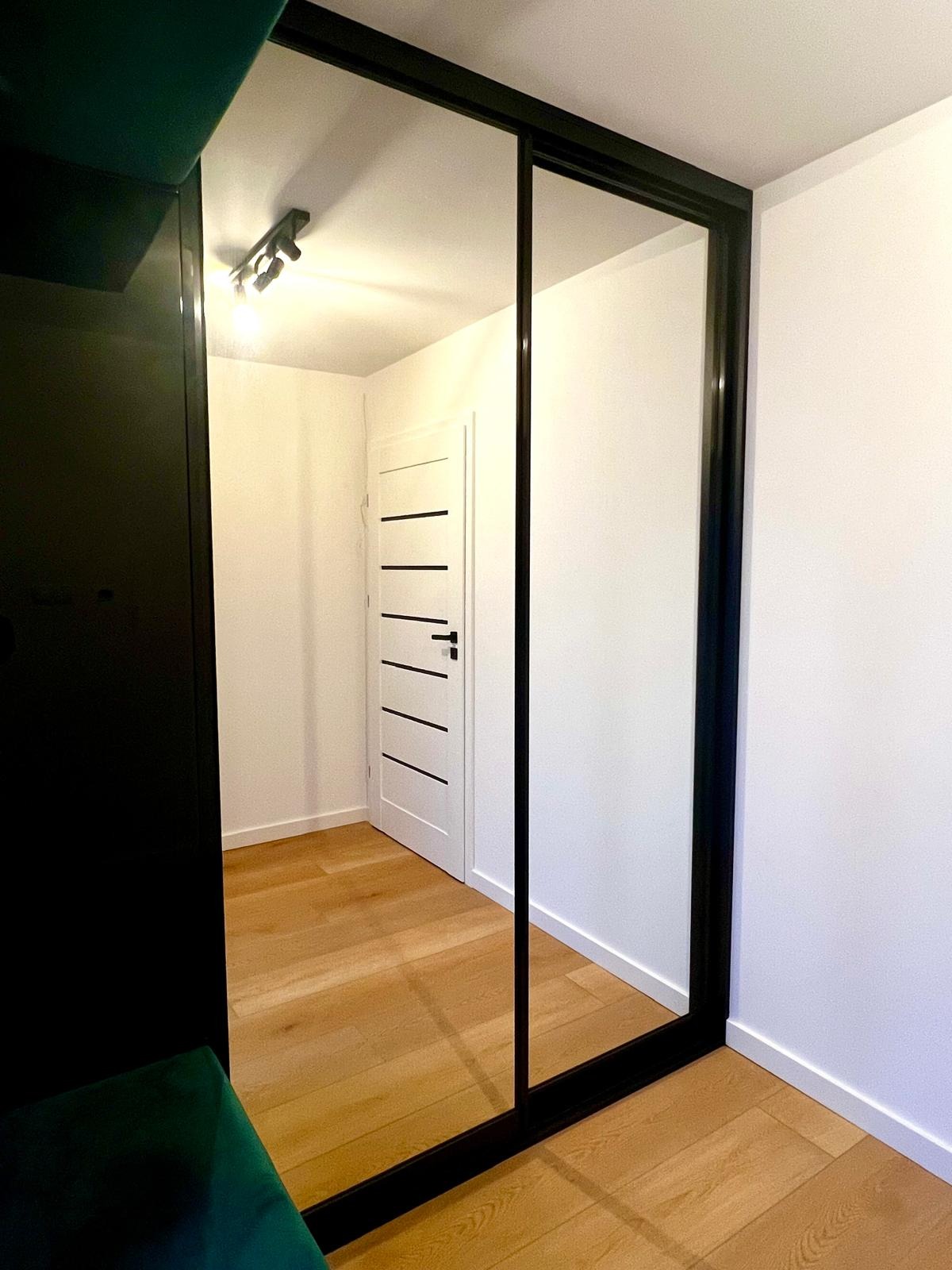 Szafa przesuwna z lustrzanymi drzwiami w czarnej ramie, odbicie wnętrza przedpokoju z białą ścianą, drzwiami z poziomymi czarnymi paskami i drewnianą podłogą.