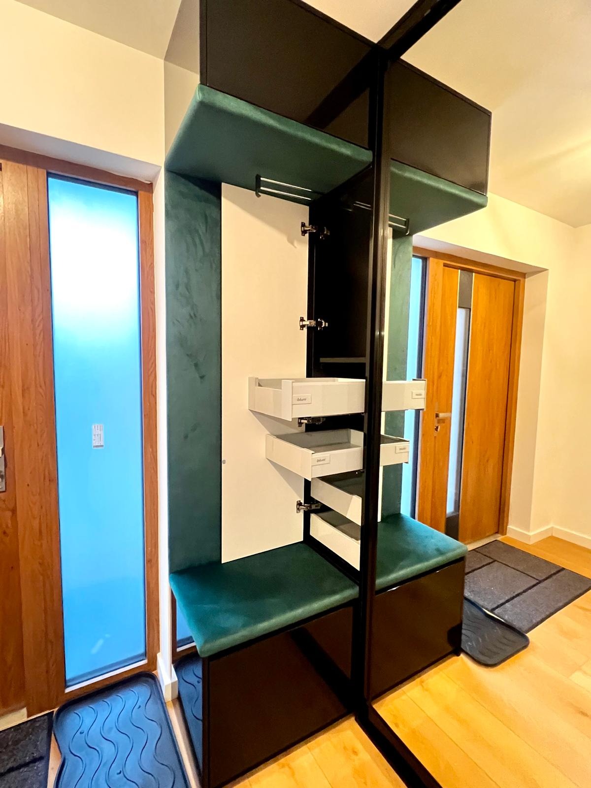 Nowoczesna szafa wnękowa z siedziskiem obitym zielonym welurem, otwarte białe szuflady z systemem Blum, czarna rama, widok na drzwi z niebieską szybą i drewniane drzwi wejściowe.