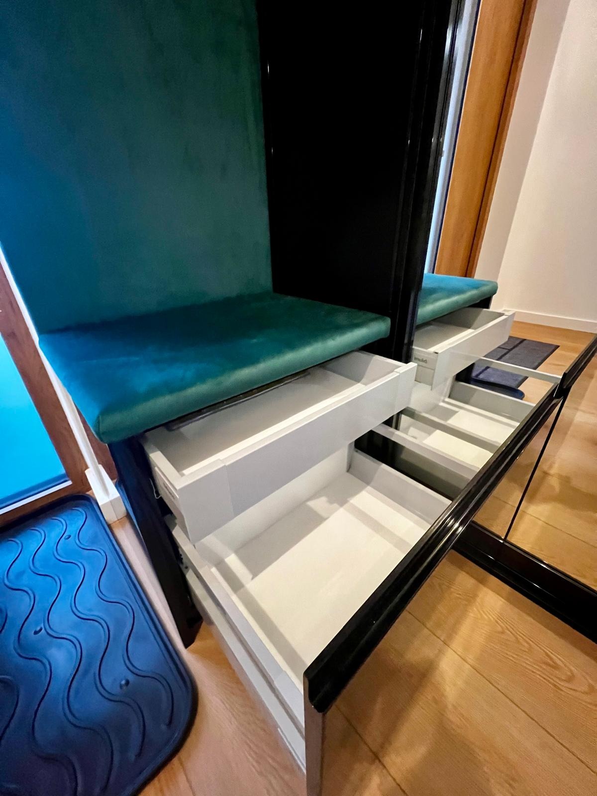 Wnętrze szafy wnękowej z siedziskiem obitym turkusowym welurem i wysuwanymi, białymi szufladami na drewnianej podłodze.