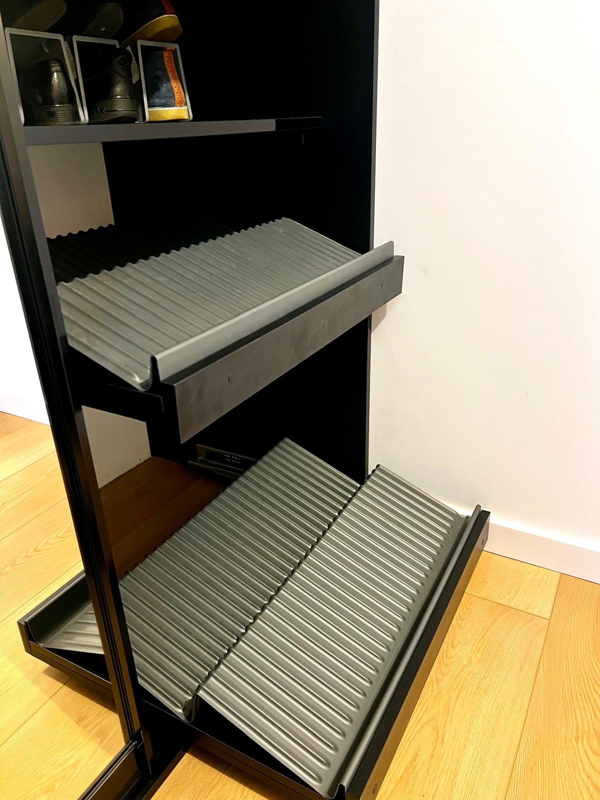 Czarna szafka na buty z wysuwanymi, żebrowanymi półkami, ustawiona na drewnianej podłodze przy białej ścianie.