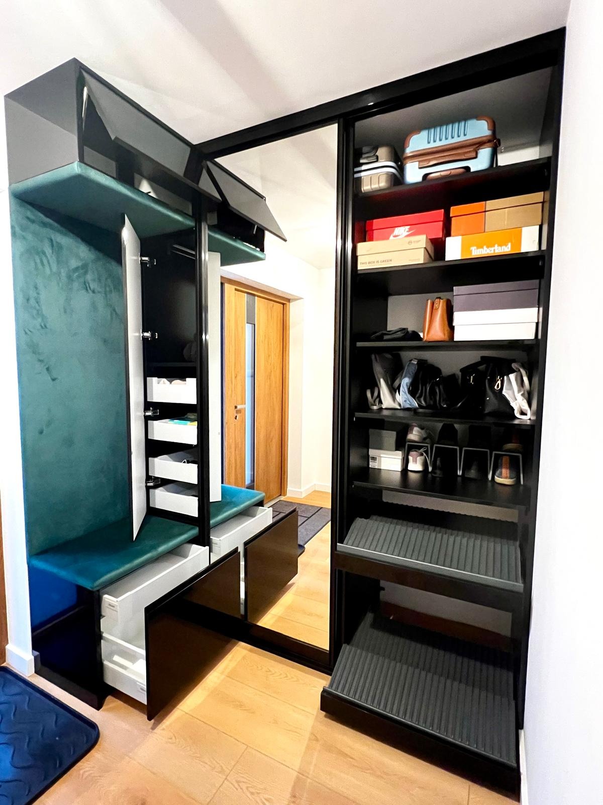 Czarna szafa wnękowa z lustrzanymi drzwiami przesuwnymi, wewnętrzne półki z pudełkami na buty i bagażem, obok siedzisko z zieloną tapicerką i otwartymi szafkami, drewniana podłoga.