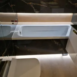 Nowoczesna łazienka z wanną, czarnymi płytkami imitującymi marmur z jasnymi żyłami, podświetleniem LED i jasną drewnianą podłogą.