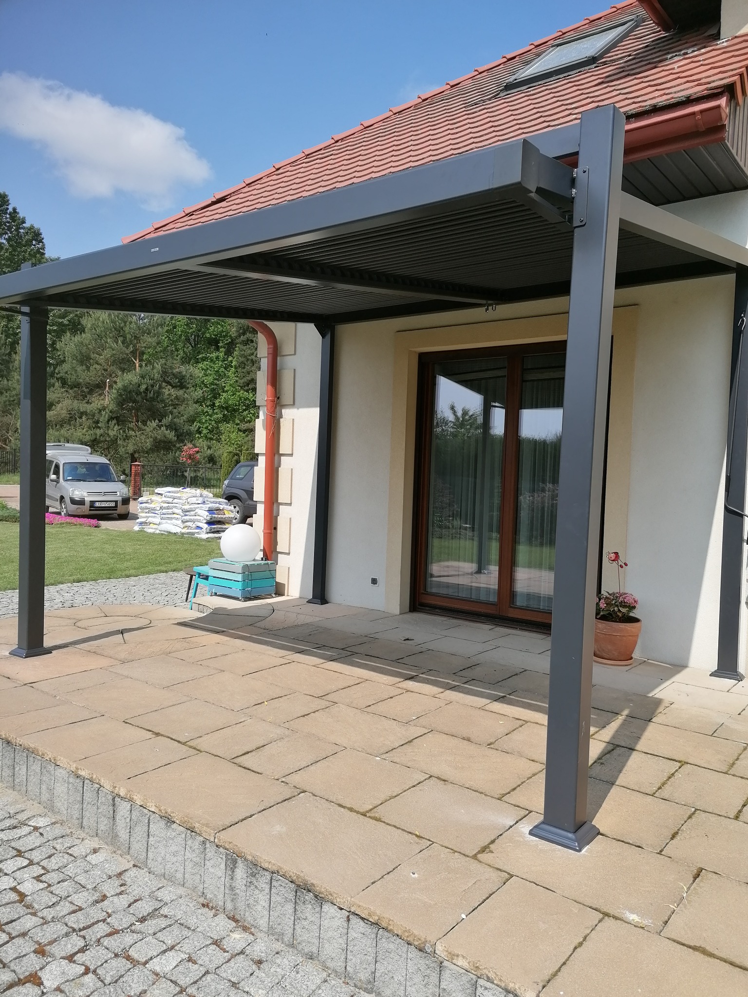Nowoczesna, grafitowa pergola lamelowa przylegająca do domu z jasną elewacją. Widoczne przesuwne drzwi tarasowe, donica z kwiatami, kostka brukowa na tarasie, trawnik, zaparkowane samochody...