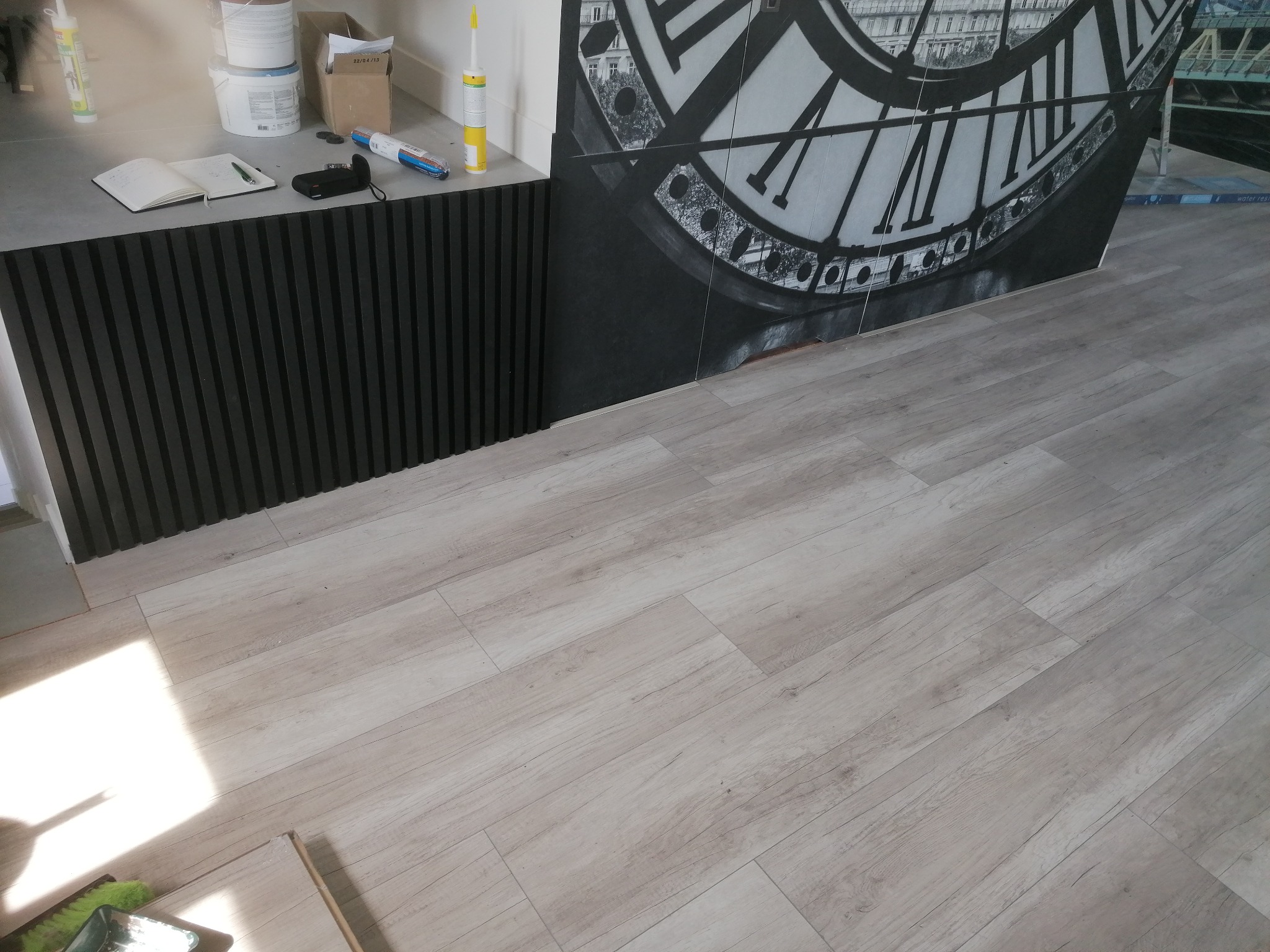 Wykończona podłoga z paneli imitujących drewno, widoczna część ściany z czarnymi listwami oraz fragment dekoracji ściennej z motywem zegara i elementami architektonicznymi w stylu retro.