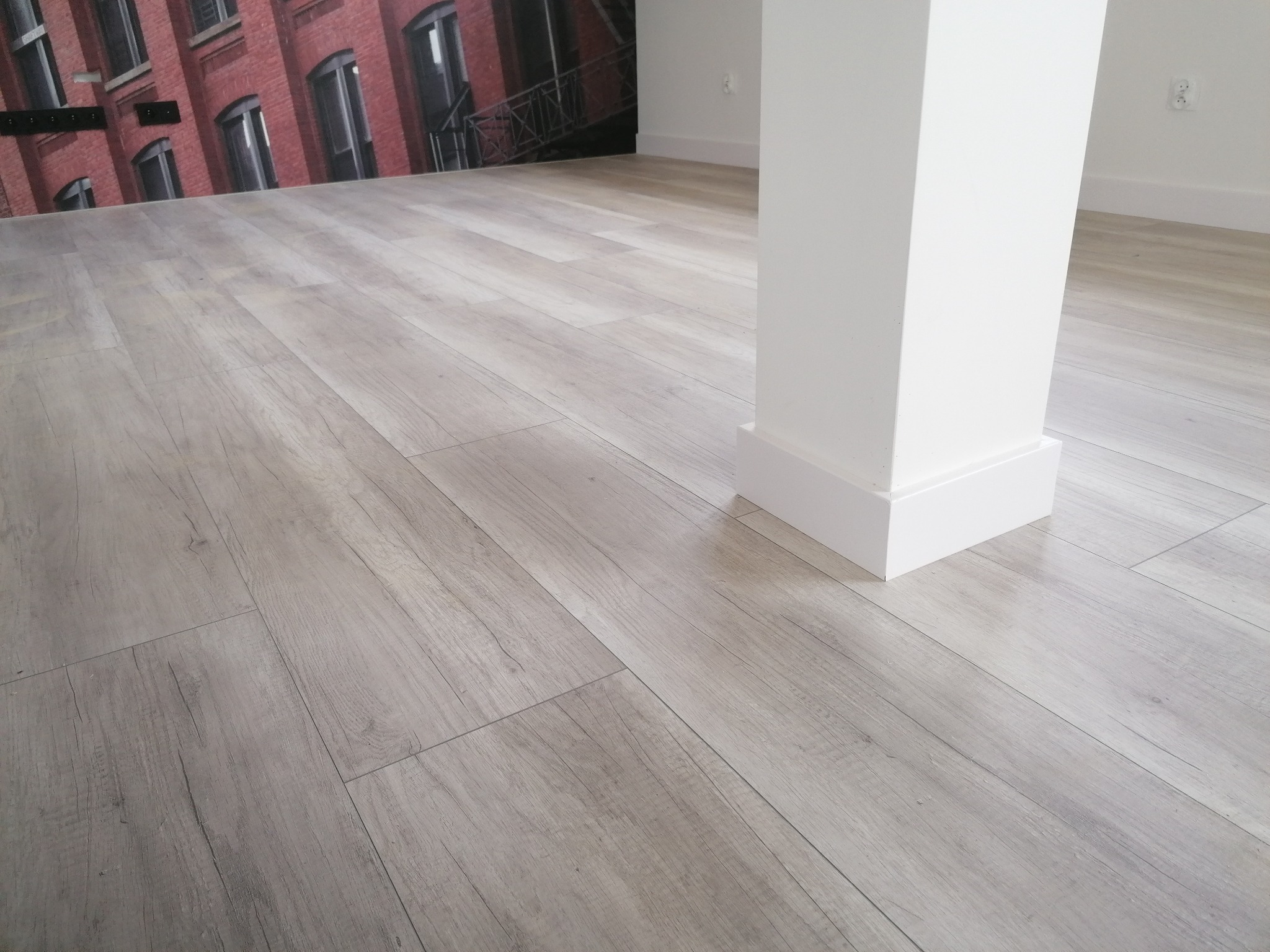 Jasnoszara podłoga z paneli imitujących drewno, ułożona wzdłużnie, z widocznym białym filarem i fragmentem ściany z fototapetą przedstawiającą ceglany budynek.
