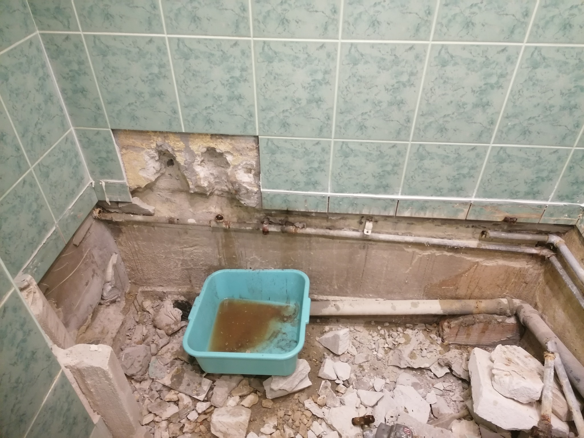 Uszkodzona ściana z brakującymi płytkami w łazience, widoczne rury, gruz i niebieska miska z brudną wodą.