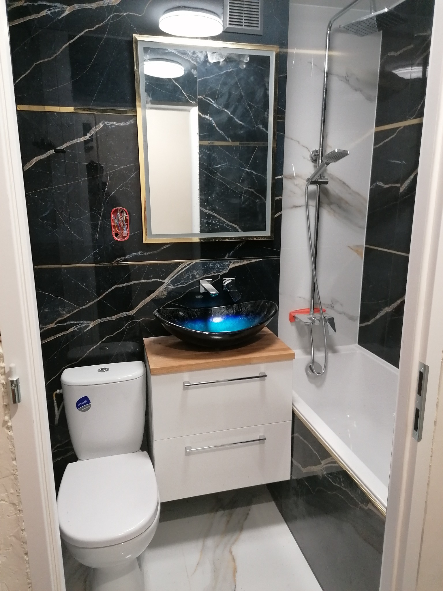 Nowoczesna łazienka z czarno-białymi marmurkowymi płytkami, umywalką w kształcie misy z podświetleniem LED, lustrem z oświetleniem i białą toaletą.