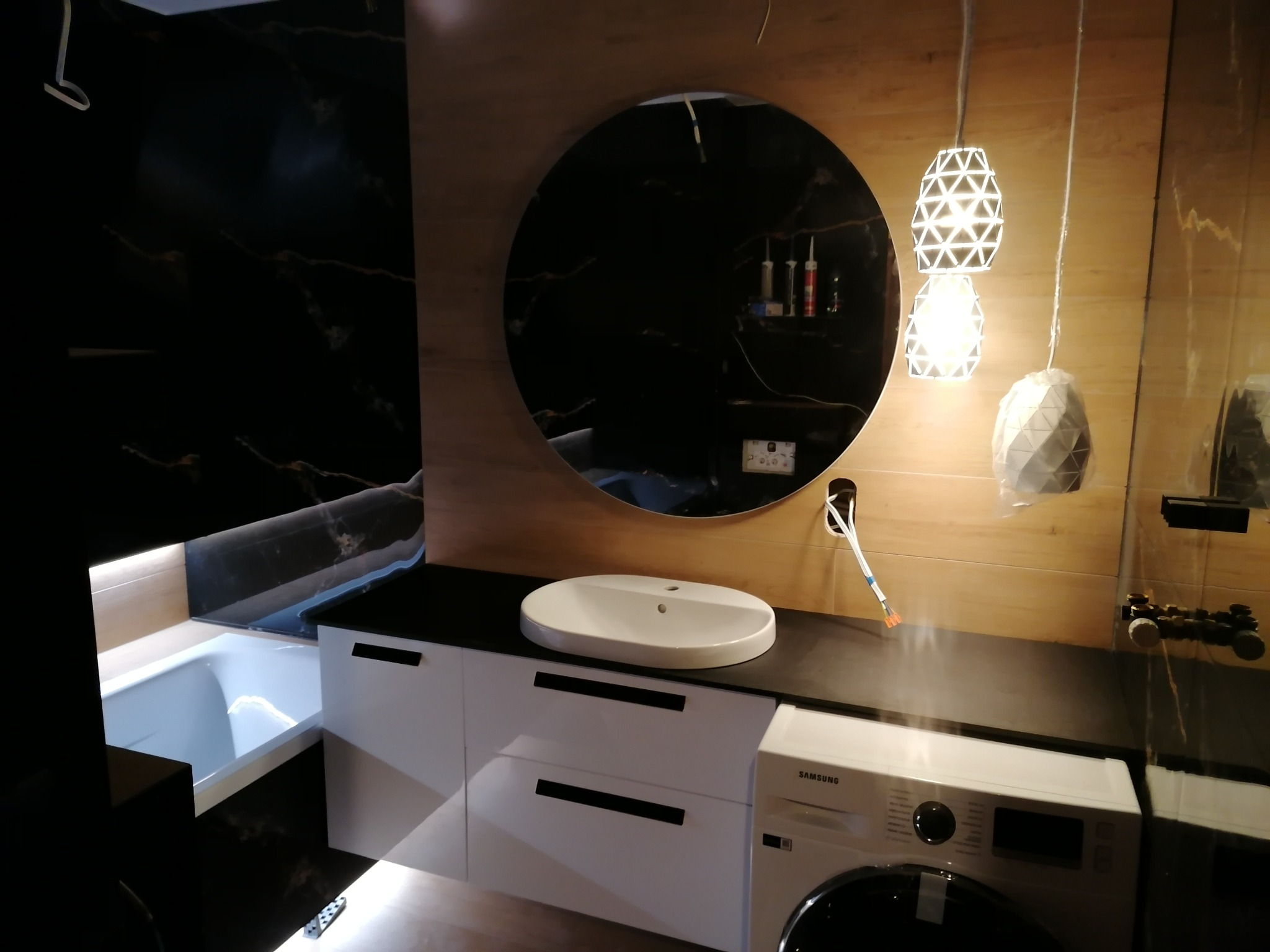 Nowoczesna łazienka z okrągłym lustrem, umywalką nablatową, białymi szafkami z czarnymi uchwytami, pralką Samsung oraz wiszącymi lampami dekoracyjnymi.