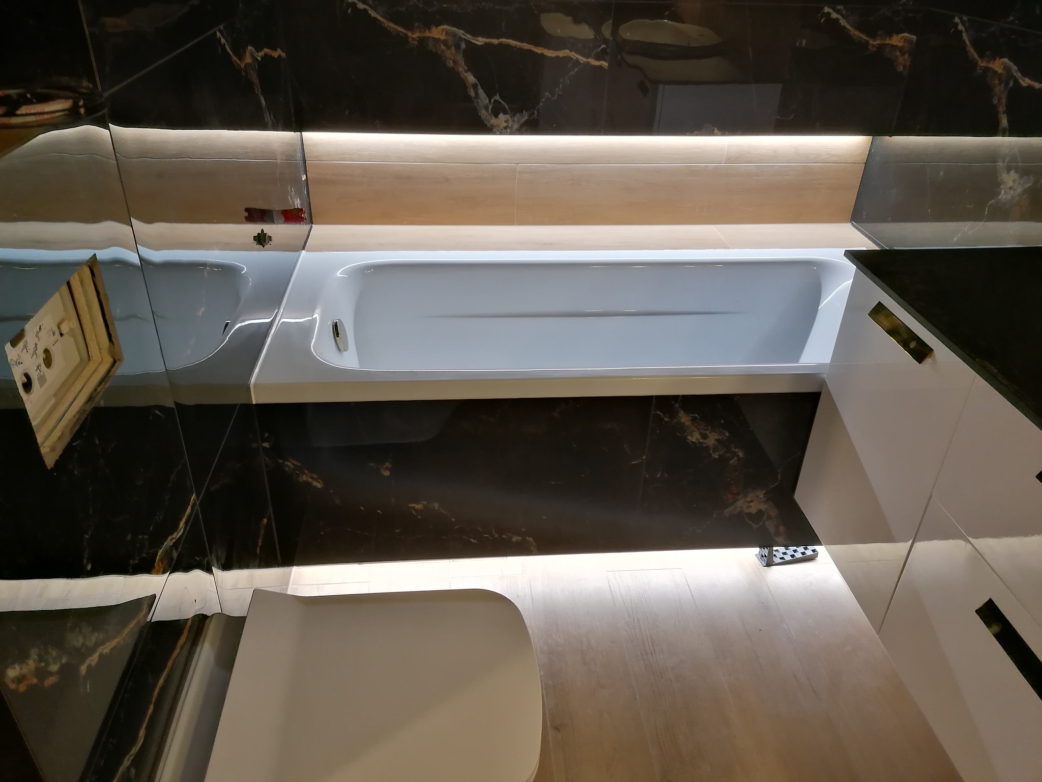 Nowoczesna łazienka z wanną, czarnymi płytkami imitującymi marmur z jasnymi żyłami, podświetleniem LED i jasną drewnianą podłogą.