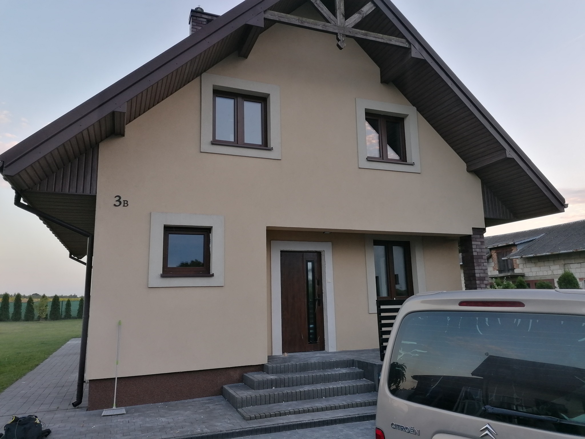 Dwukondygnacyjny dom z beżową elewacją, brązową stolarką okienną i dachem, widoczny z przodu, z samochodem zaparkowanym przed wejściem, numer domu 3B na ścianie.