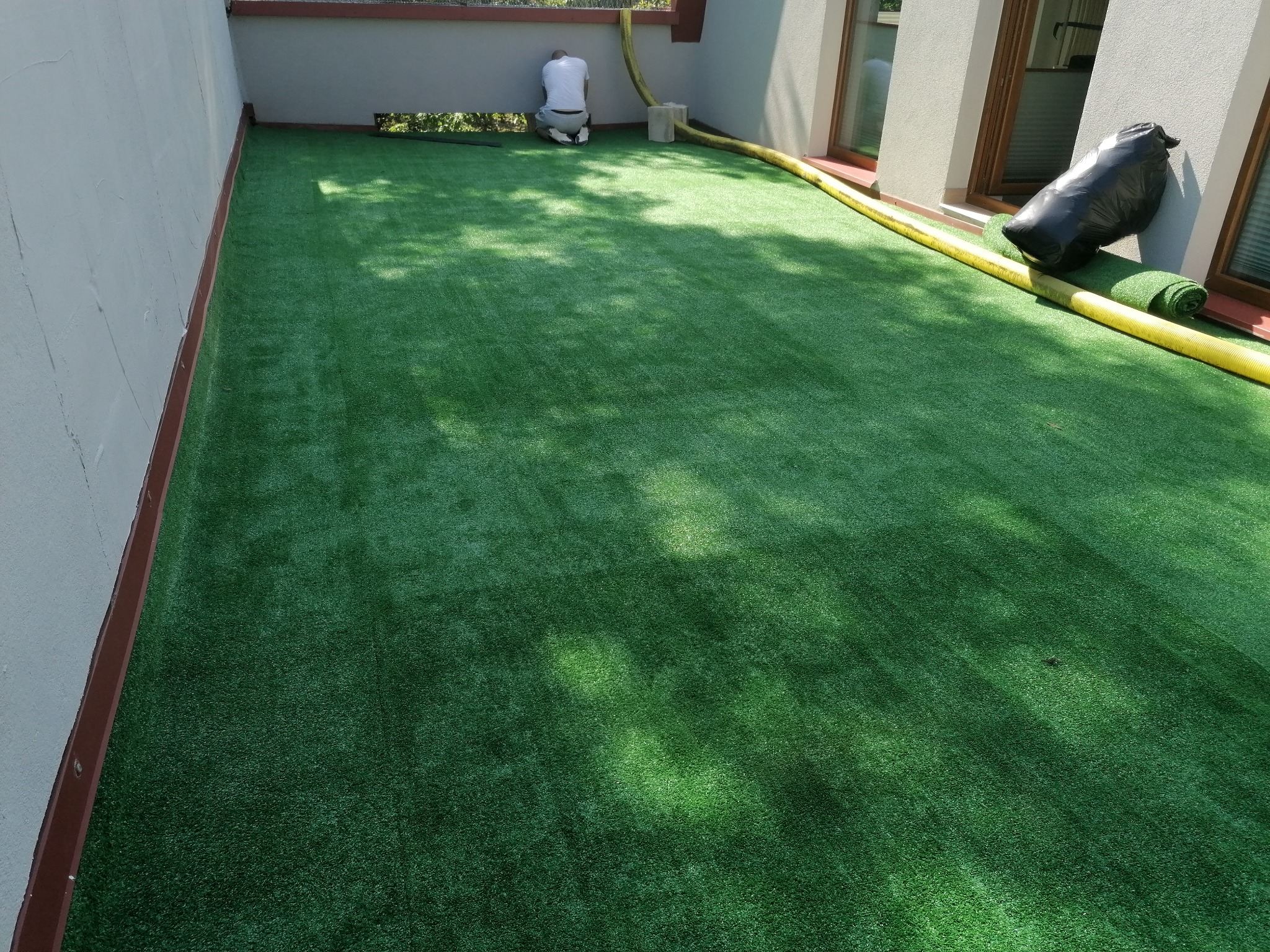 Instalacja sztucznej trawy na balkonie: mężczyzna w białej koszulce i jasnych spodniach klęczy przy ścianie, układając zieloną trawę. Obok leżą zwinięte rolki trawy i żółty wąż. W tle widoczne okna...