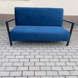 Fuerte - Dwuosobowa sofa obita niebieskim welurem, z czarnymi metalowymi nóżkami, stojąca na brukowanym chodniku.
